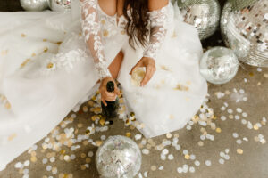 bride in a white wedding gown sitting on the floor surrounded by confetti and disco balls, drinking champagne 