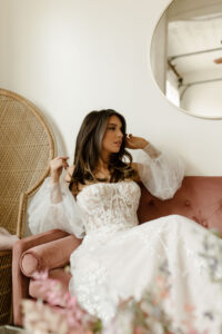 relaxed bride to be wearing a lace off the shoulder wedding dress while sitting on a pink couch in Ivoryology bridal store near 