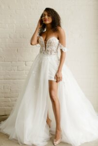 stunning bride with curly dark hair wearing an off the shoulder wedding dress with a lace bodice and a short hemline, with a unique floor length skirt in front of a white brick wall at Ivoryology in the Memphis area