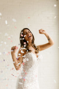 happy bride with arm tattoos in white lace form fitting wedding dress with white and pink confetti falling around her