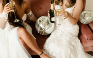 two brides in white wedding dresses sitting on a pink velvet sofa, surrounded by disco balls, drinking champagne ready to toast their decision to buy their bridal gowns at Ivoryology in Memphis