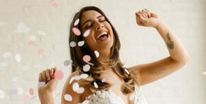 A happy bride dancing through a cloud of confetti while wearing a sleeveless lace wedding dress.