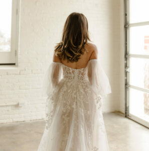 A bride with her back to the camera, modeling an off the shoulder long sleeve wedding dress.