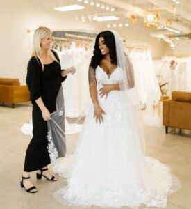 Smiling bride to be wearing a stunning wedding dress and veil and standing with her wedding dress consultant.