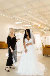 Smiling bride to be wearing a wedding dress and veil and standing by her wedding dress consultant.